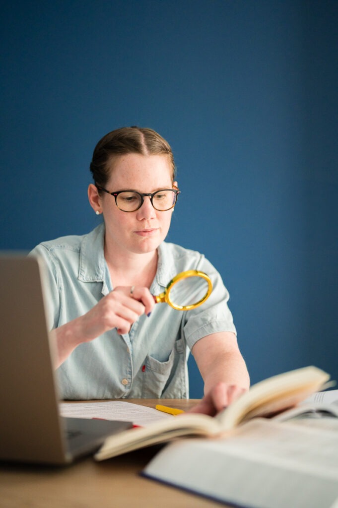 Philippine SIKORA, copy editor and proofreader, hold a magnifying glass in her right hand and books in her left hand.