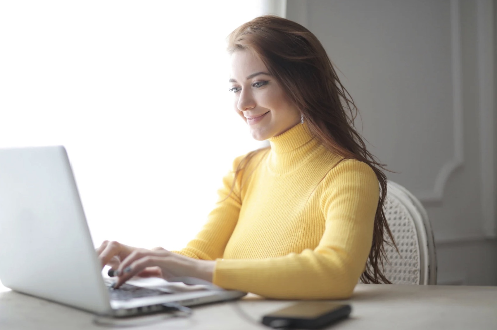 A smiling woman wearing a yellow jumper working on her laptop.